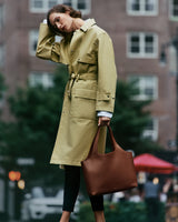 Person wearing a trench coat holding a large tote bag standing on a city street.