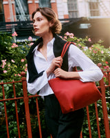 Person with a cape over shoulders holding a travel bag, standing near a fence with flowers in background
