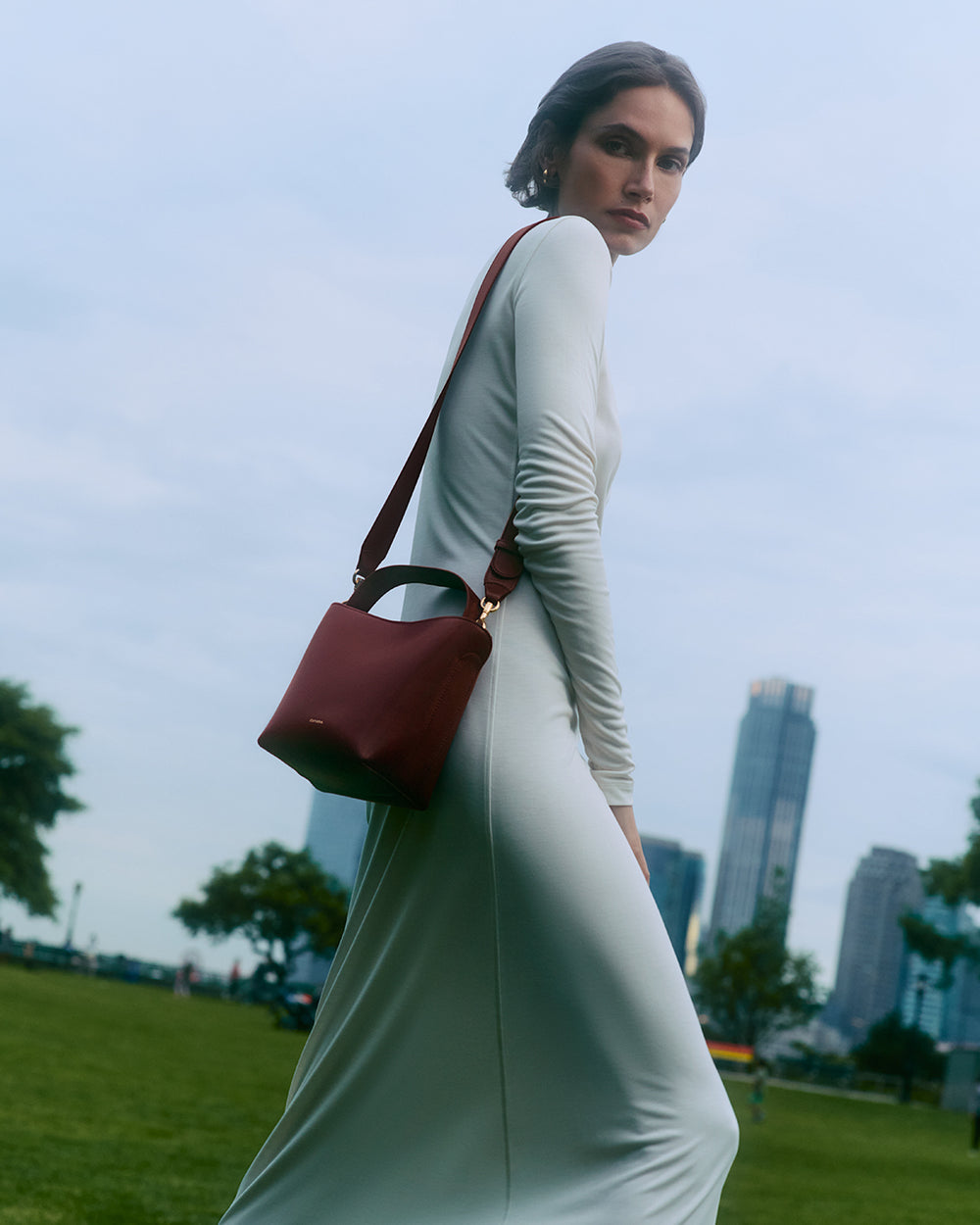 Mini Linea Bucket Bag Woman in a long dress standing outside with a shoulder bag, city skyline in the background