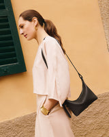 Woman in profile with a crossbody bag, standing near a wall and window