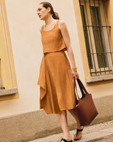 A person in a loose sleeveless top, and flowy skirt holds a large handbag walking by a building.