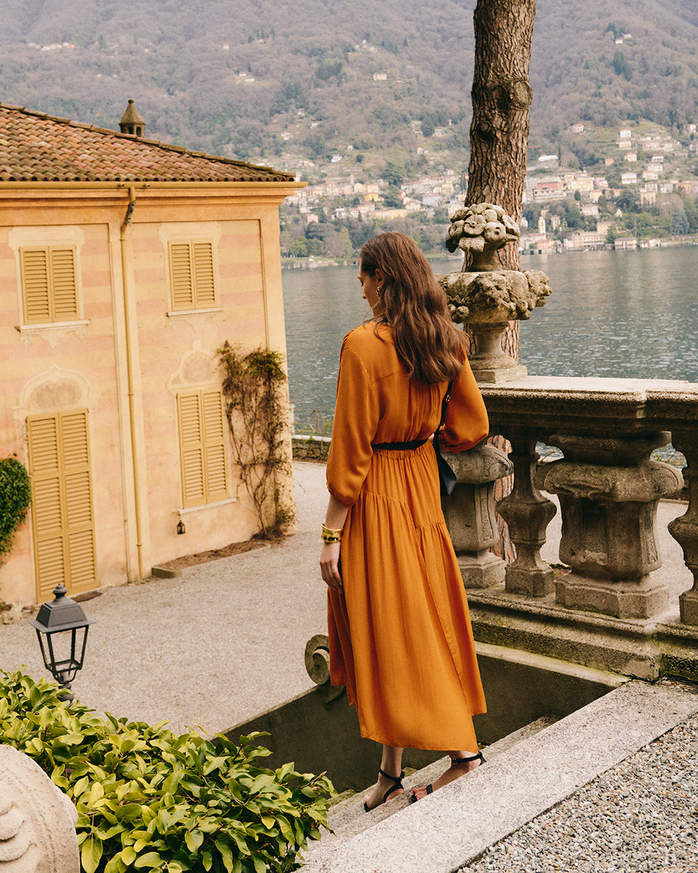Person in long dress stands by stone railing overlooking water and hillside town.