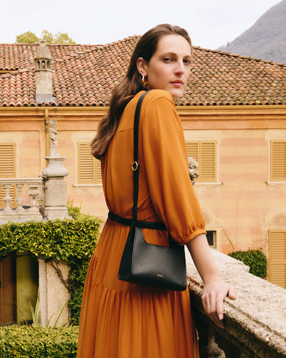 Woman standing on a balcony with a shoulder bag, looking at the camera, building in background.
