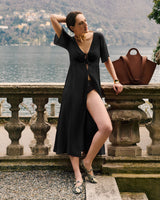 Person in dress leans on balcony with bag, scenic lake and mountains in background.