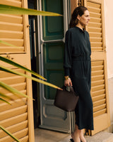 Woman in long attire holds bag standing by open shutters.