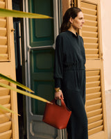 Person in a jumpsuit holding a handbag standing by wooden shutters.