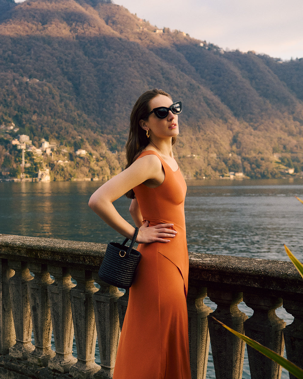 Woman in dress by a stone railing with a woven bucket bag, lake and mountains in background.
