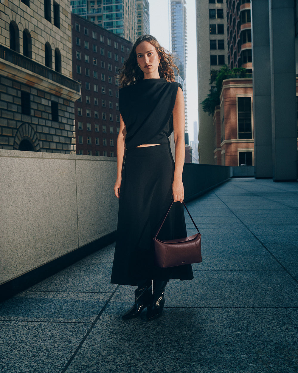 Person in a sleeveless top and long skirt holding handbag, standing on city street between tall buildings.