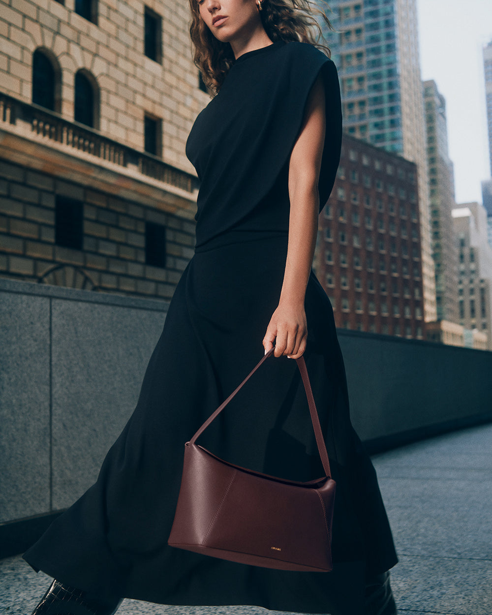 Person in flowing sleeveless top and long skirt, holding a handbag, urban buildings in background.