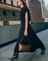 Person walking on a city street holding a handbag wearing a long skirt and boots.