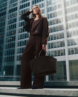 Woman in suit stands on steps holding large bag with city buildings in background.