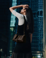 Woman standing outdoors with handbag, looking upwards among tall buildings.
