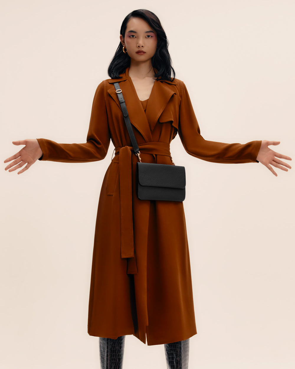 Person wearing a long coat and a shoulder bag, standing with arms slightly outstretched.