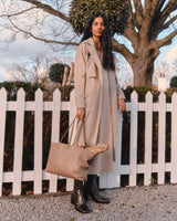 Woman standing by a white picket fence with a tree in the background, holding a bag and wearing boots.