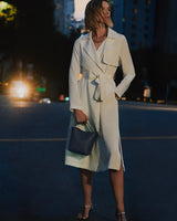 Person in a long coat holding a handbag while standing on a city street.