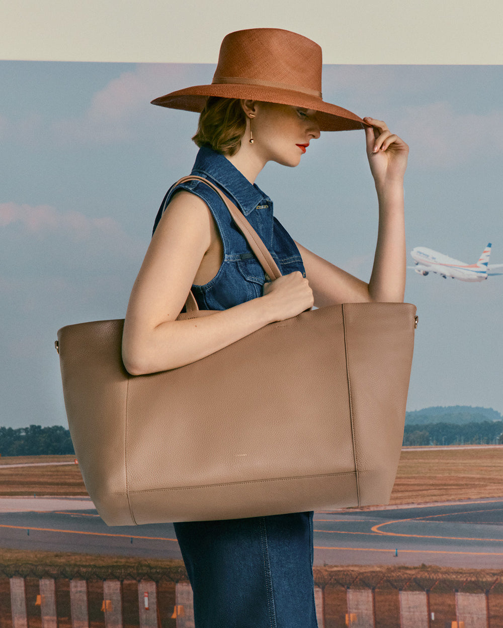 Woman holding large bag, touching hat brim, airplane in background at airport.