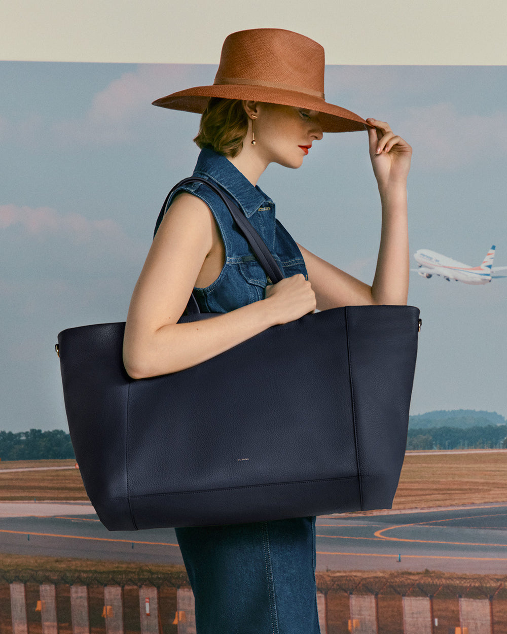 Woman holding large bag and adjusting wide-brimmed hat with airplane in background.