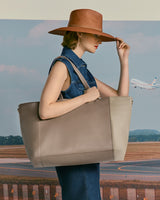 Woman holding a large bag, adjusting wide-brimmed hat, with an airplane in the background.