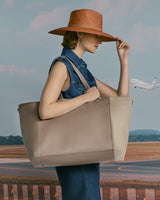 Person holding a large bag standing in front of a runway with a plane in the background, wearing a hat.