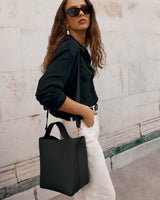 Person wearing sunglasses with a shoulder bag, blouse, and pants, standing against a stone wall