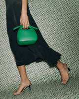 Woman in a dress and sandals holding a handbag, walking in front of patterned wall.