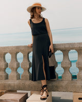 Woman in dress and hat standing by stone railing near the sea, holding a small bag.