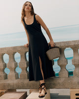 Woman in sleeveless long dress holding woven bag, standing by stone balustrade overlooking the sea.
