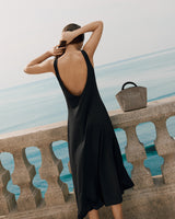 Woman in dress standing by stone railing with view of ocean, holding her hair up. Handbag on the railing