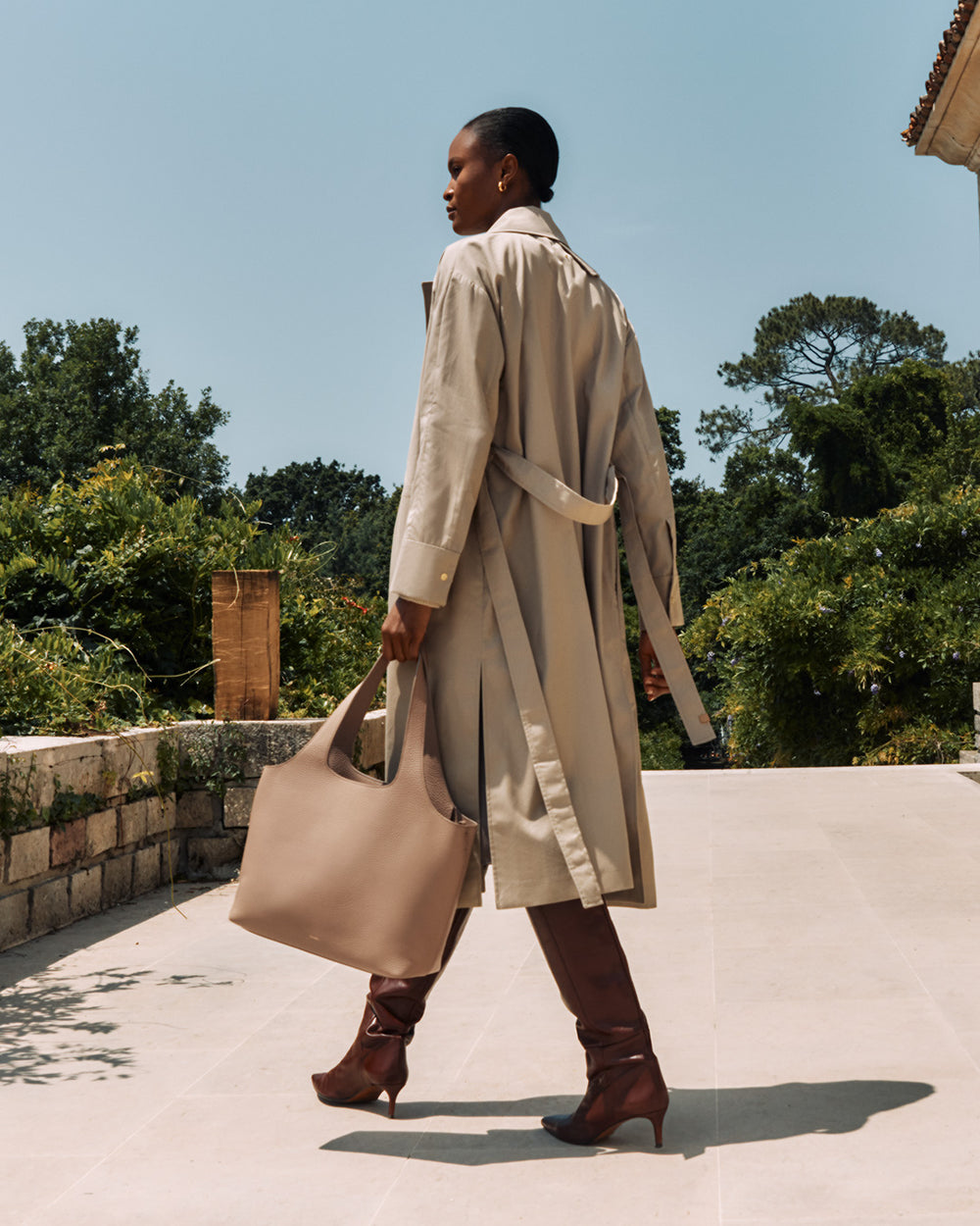 Woman walking outdoors wearing a long coat and holding a large bag.