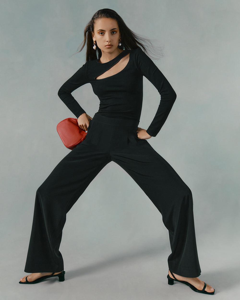 Woman standing in a wide stance with one hand on hip and holding a bag.
