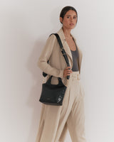 Woman standing with a handbag, wearing a blazer and trousers.