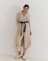 Woman standing in a long coat with a belt, wearing loafers, and looking at the camera.