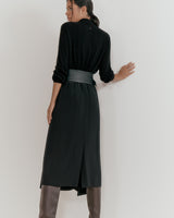 Woman standing in a mid-length dress and boots, viewed from the back, leaning on a wall.