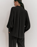 Woman seen from behind wearing a blazer and pants standing against a plain background.