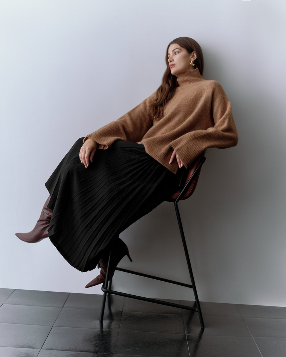 Person sitting on a chair wearing a loose sweater and a pleated skirt with boots.