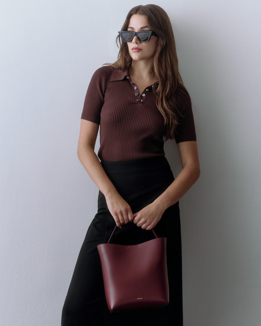 Linea Bucket Bag Woman in sunglasses holding a handbag, wearing a short-sleeve collared top and skirt.