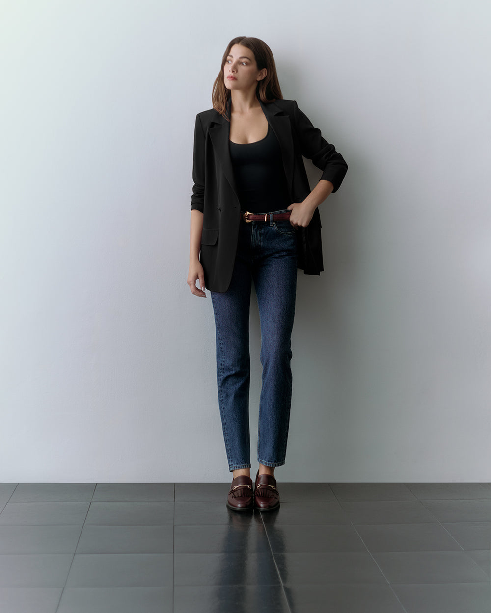 Person standing against a wall wearing a black blazer, black top, jeans, and loafers.