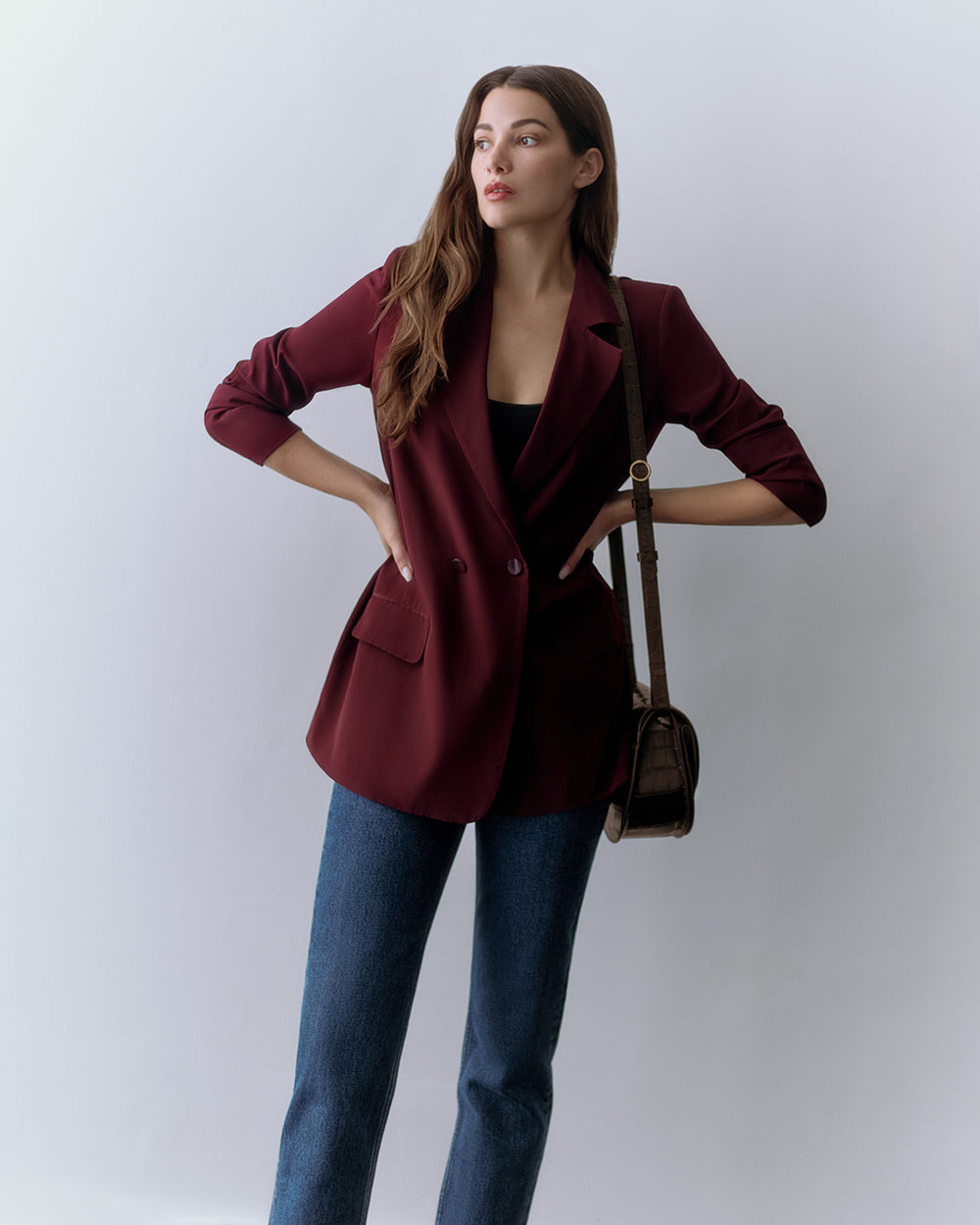 Person wearing a red blazer, top, jeans, and croc-embossed leather shoulder bag, standing with hands on hips