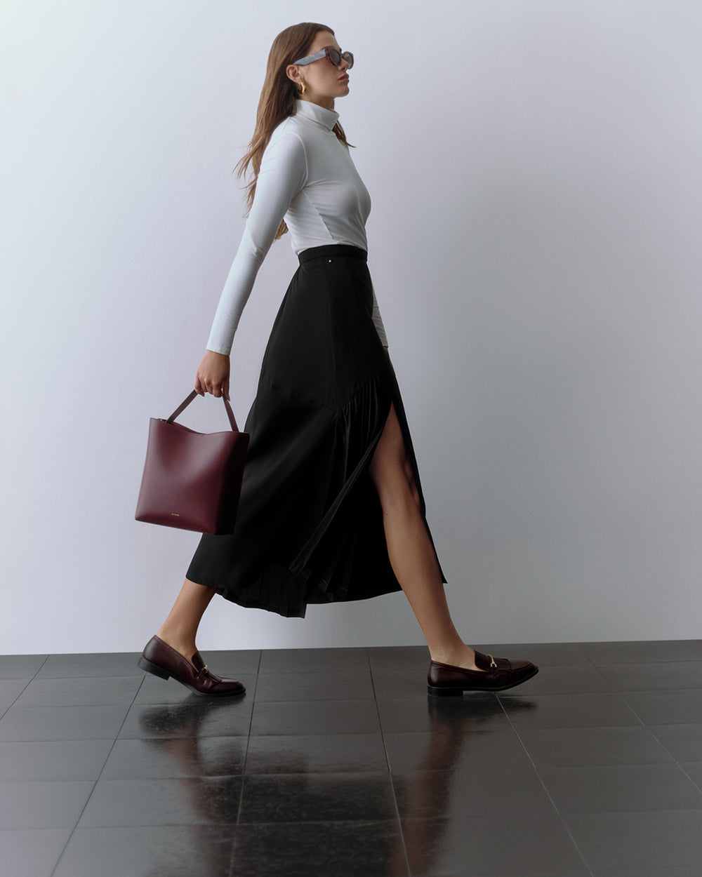 Person walking, wearing a fitted top and long skirt, holding a tote bag and wearing sunglasses.