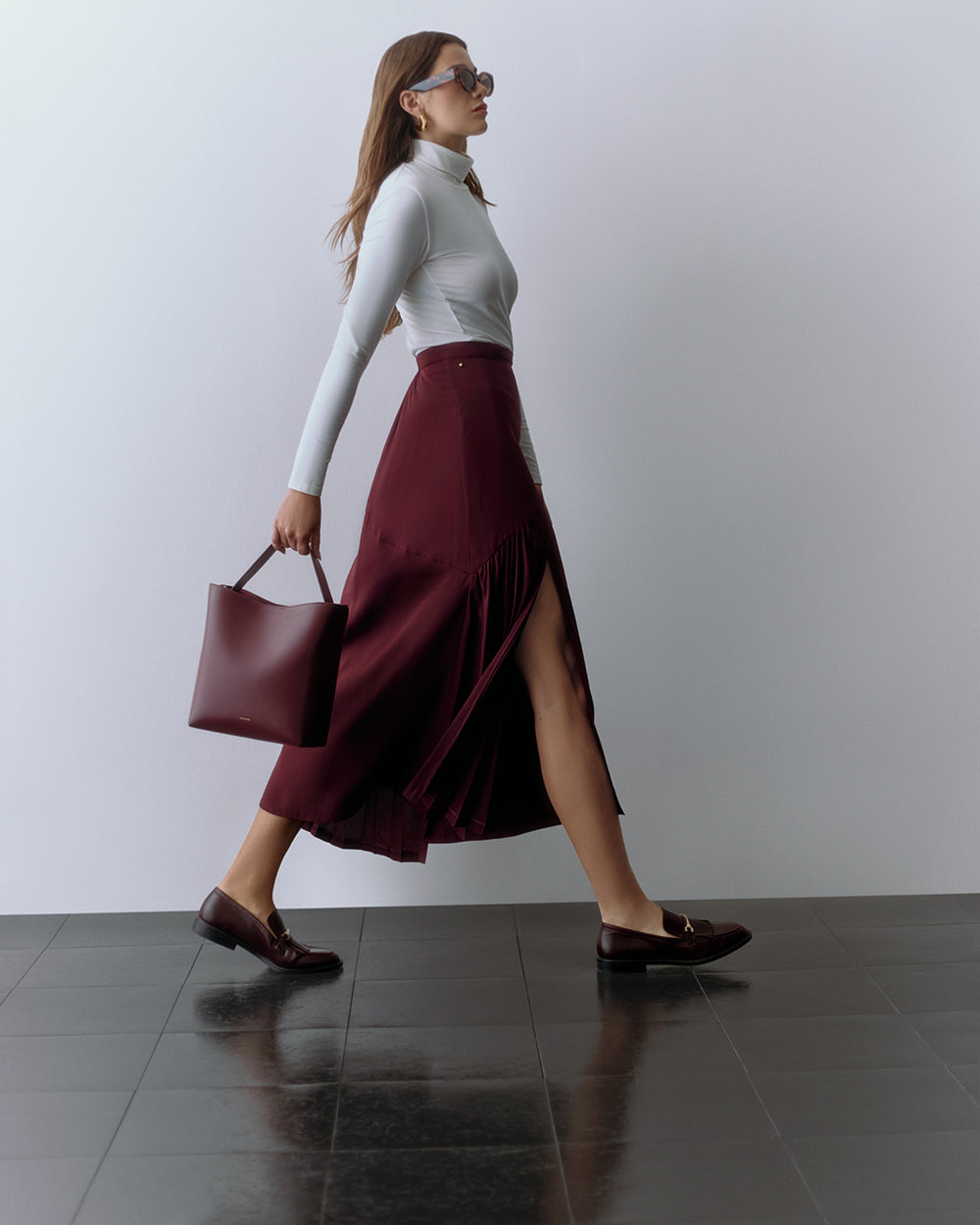 Person walking, wearing a fitted top and long skirt, holding a tote bag and wearing sunglasses.