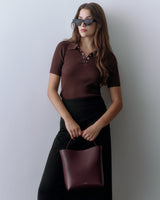 Woman with sunglasses holds a handbag, wearing a short sleeve top and long skirt, standing indoors.