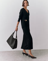 Person stands holding large handbag on their left side, wearing a long dress and heels.