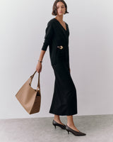 Woman in long dress holds handbag, wearing heels, standing on plain background.