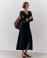 Person in long dress with large shoulder bag, arms crossed, standing indoors.