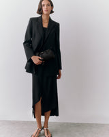 Person in jacket and dress holds a small bag, wearing sandals, standing against plain background.