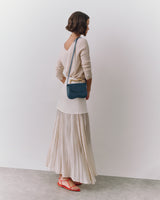 Person standing, wearing long skirt, top, holding small shoulder bag, and wearing sandals.