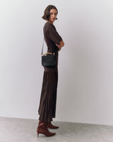 Woman in long dress with shoulder bag and boots standing indoors.