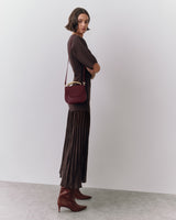 Person standing with a small shoulder bag and pleated long skirt wearing boots.