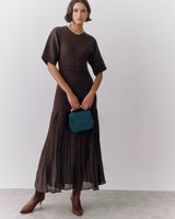 Woman in long dress holding a small handbag, wearing boots, standing in a minimalist room.