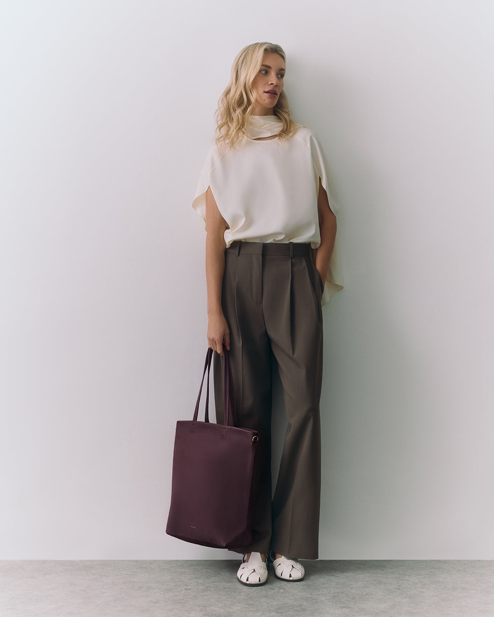 Woman in sleeveless top and trousers holding a tote bag standing against a wall
